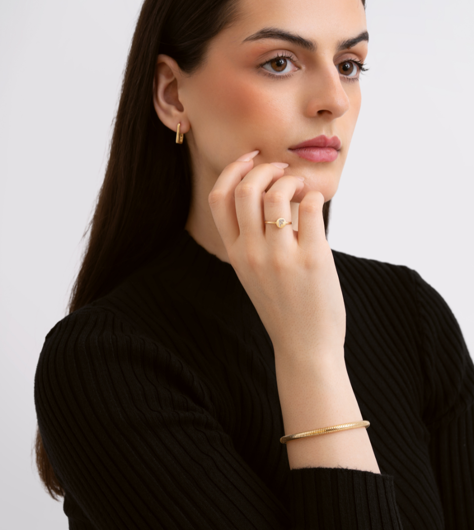 Woman wearing gold jewelry including earrings, ring, and bracelet against a plain background